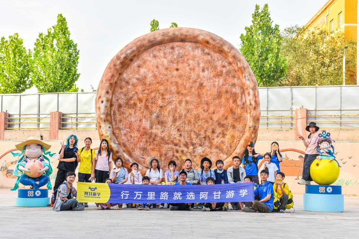 孩子们在新疆大馕雕塑前合影，拉着「行万里路 就选阿甘游学」横幅